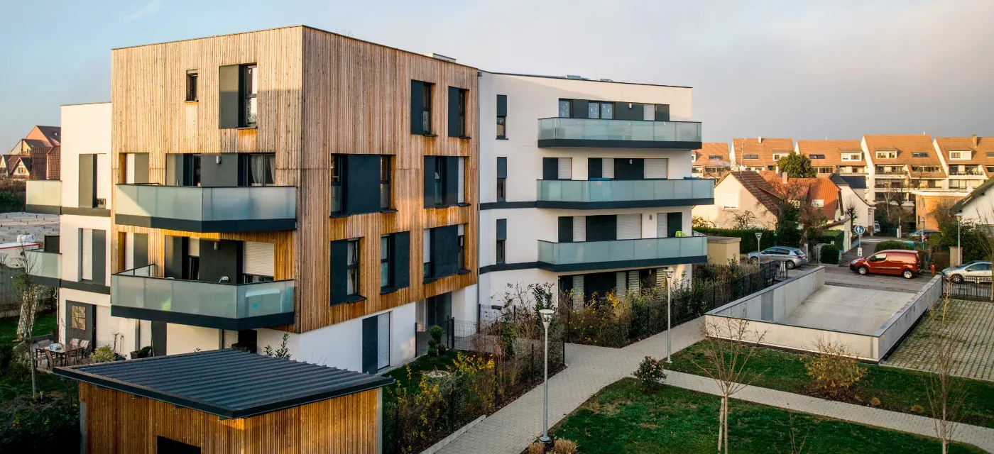 Modernes Mehrfamilienhaus mit Holzverkleidung und Glasbalkonen. Angrenzende Gebäude und Straße.