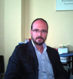 Hombre con gafas y barba, vestido con blazer, sentado en la oficina.