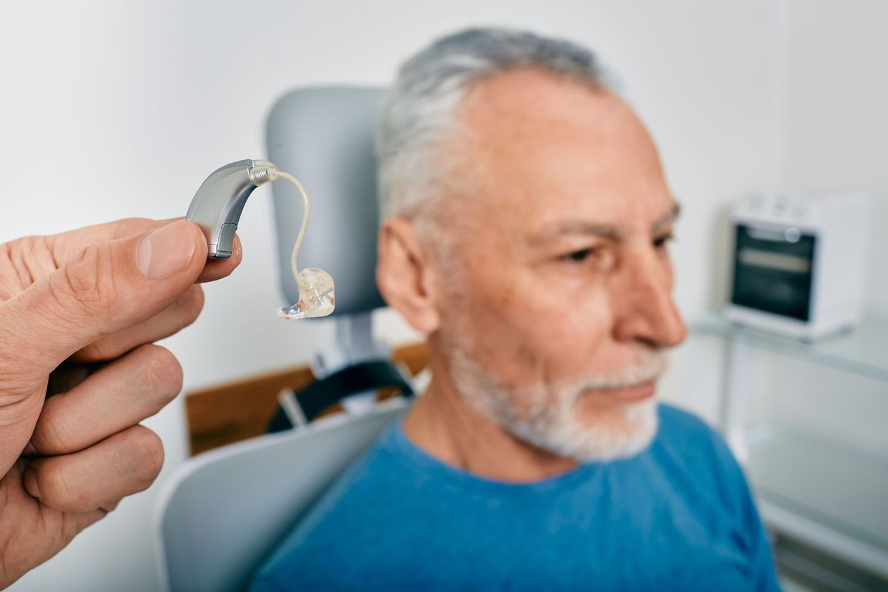 Homme âgé assis dans un fauteuil d'auscultation et audioprothésiste tenant son appareil auditif dans les mains