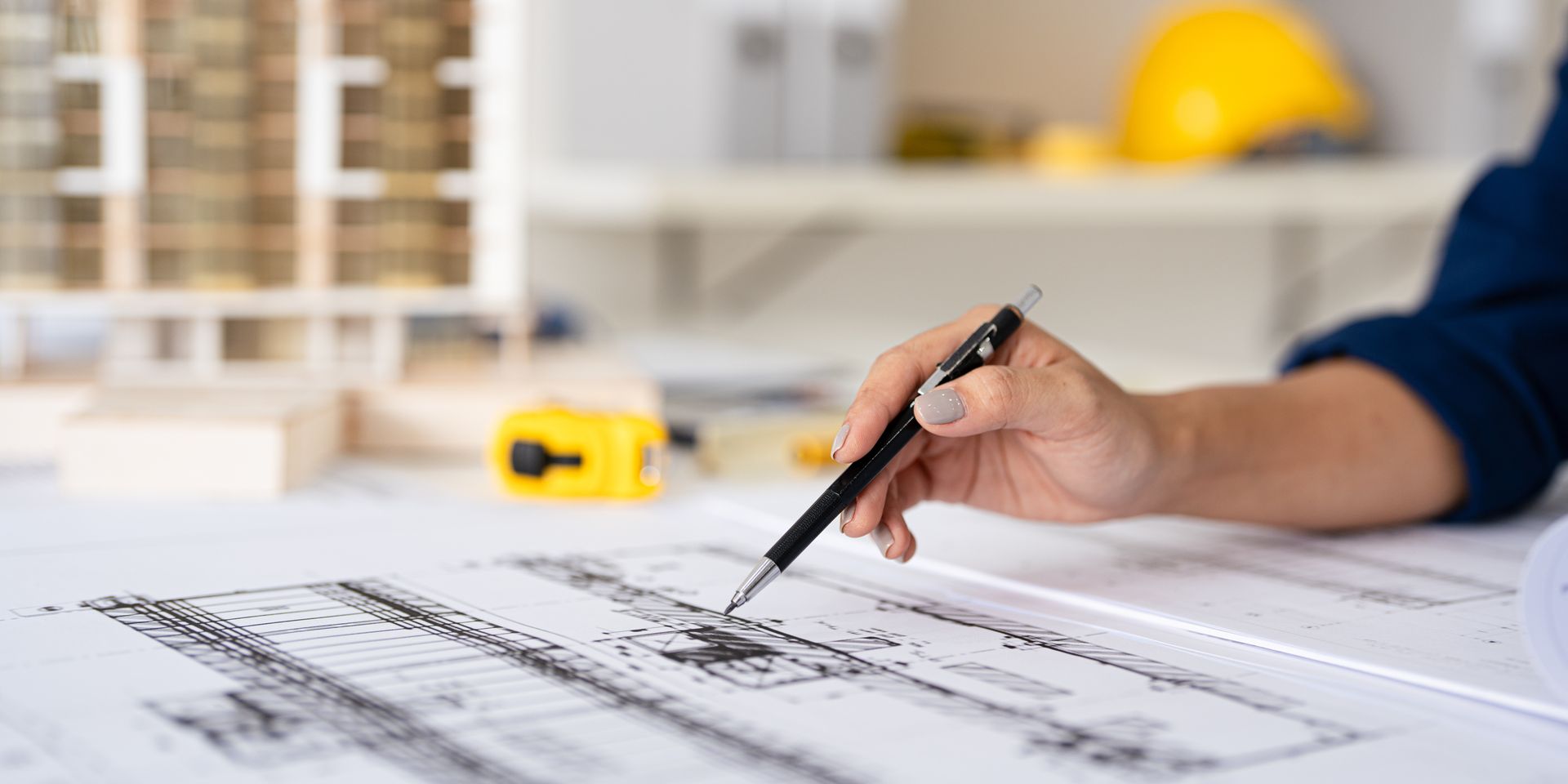 Mano che tiene una penna e indica un progetto architettonico, con un casco giallo e un modello di edificio sullo sfondo.