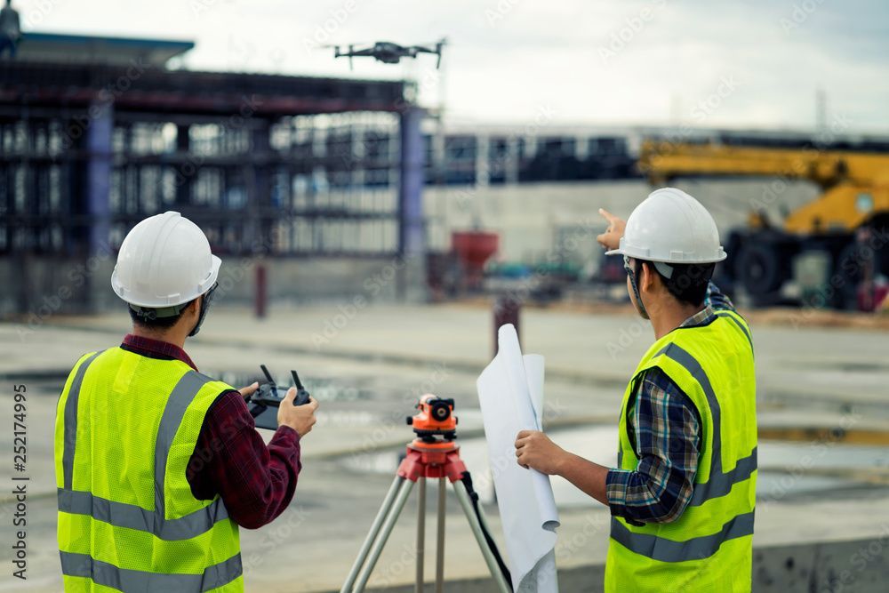Due operai edili utilizzano un drone in un cantiere. Uno punta, l'altro aziona un radiocomando.