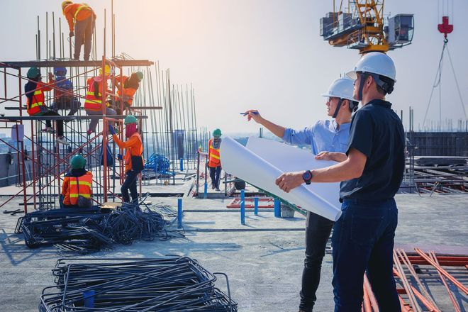 Cantiere con operai, due persone che esaminano i progetti, gru e travi d'acciaio.