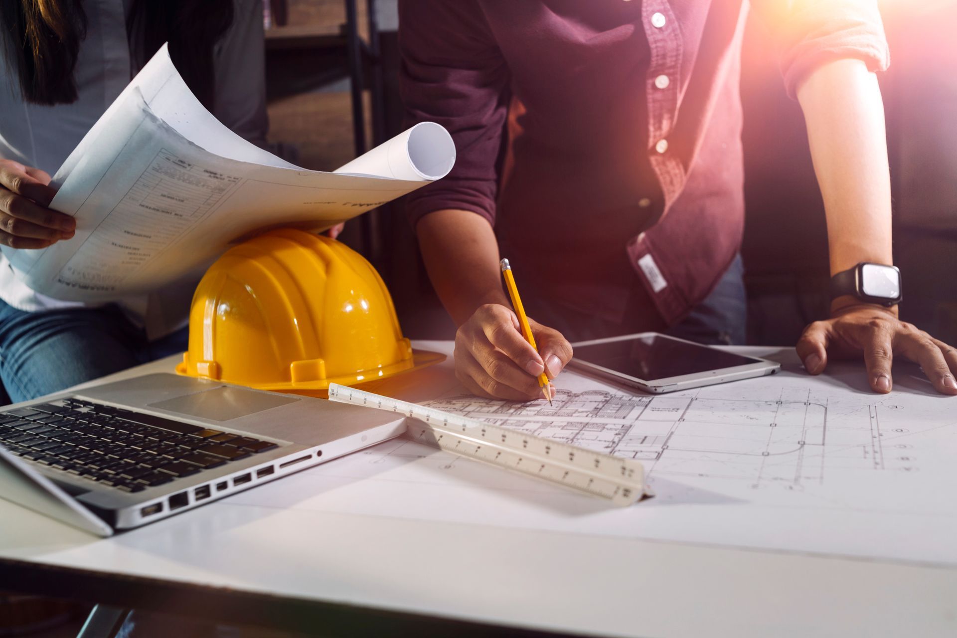 Due persone lavorano su progetti, casco giallo, computer portatile, tablet, righello e matita su un tavolo.