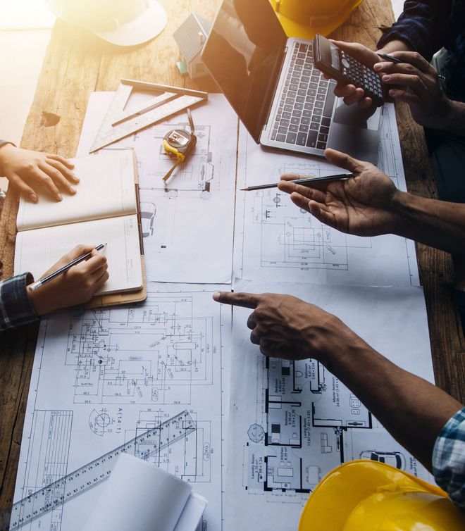 Architetti collaborano su progetti su un tavolo di legno, indicando e scrivendo, con a disposizione strumenti.