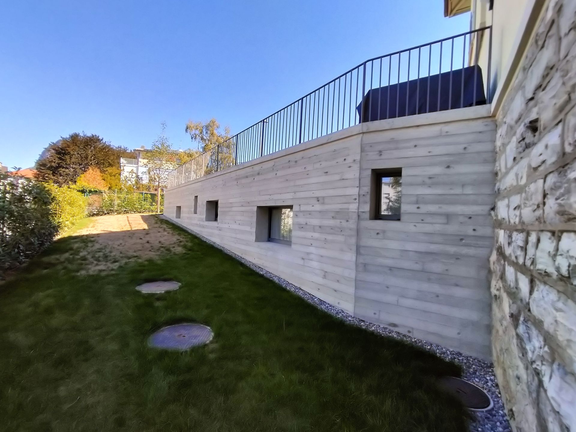 Muro di cemento con piccole finestre, balcone con ringhiera nera e prato.