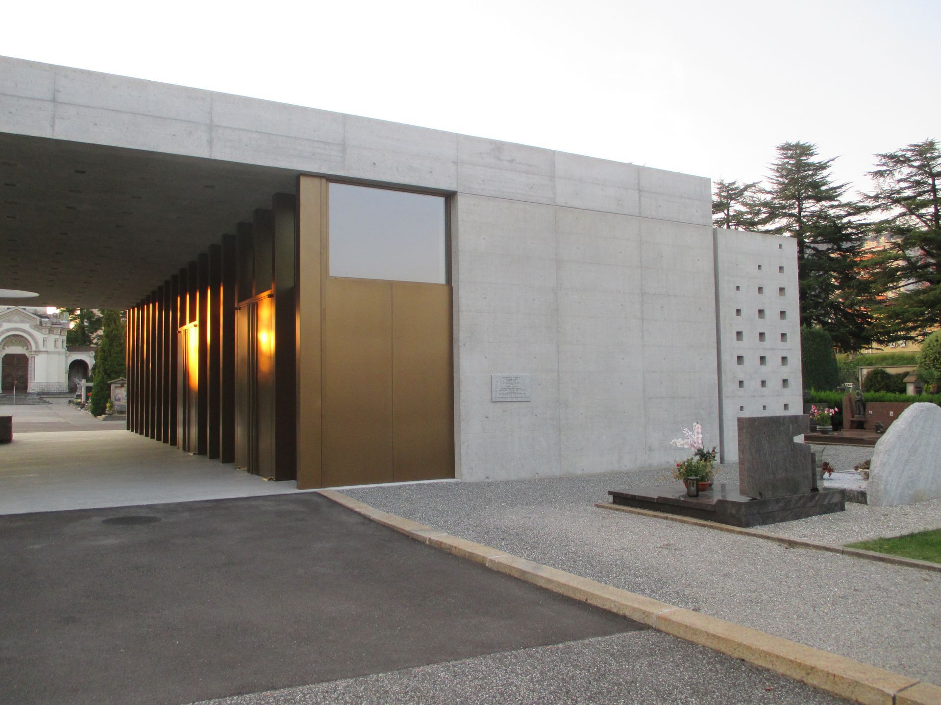 Moderno edificio in cemento con porta dorata, passaggio pedonale aperto e un cimitero con lapidi.