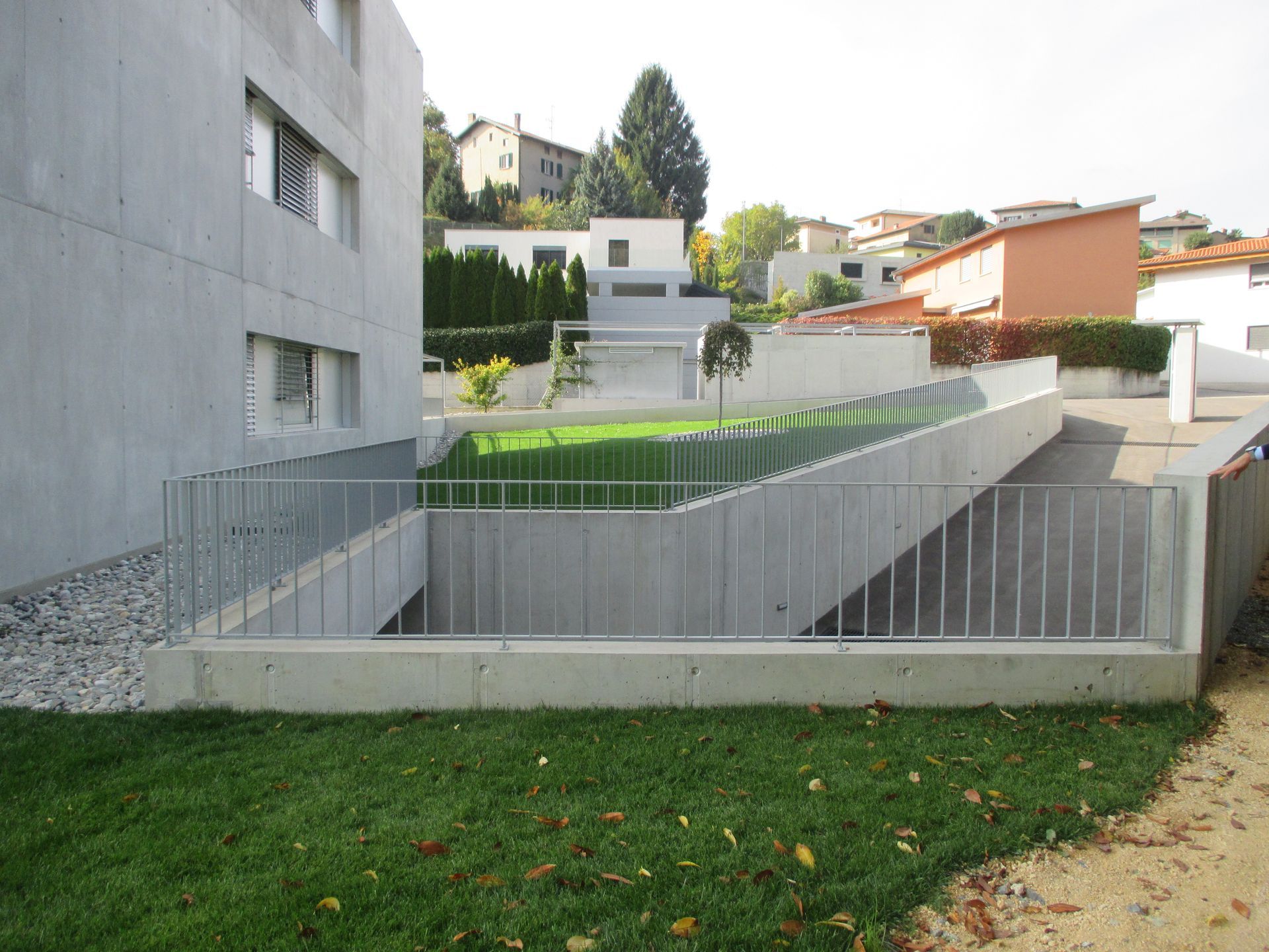 Edificio in cemento con rampa che conduce a uno spazio verde, ringhiera in metallo, zona residenziale sullo sfondo.