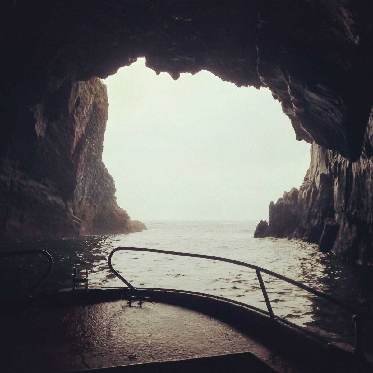 Vista desde una oscura cueva marina que se abre a un océano brumoso con una barandilla metálica mojada en primer plano.