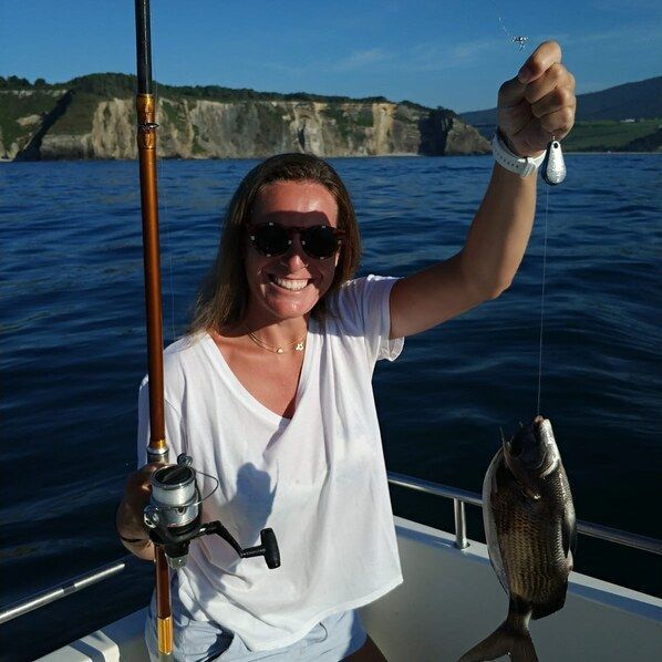 Una persona en un bote sosteniendo una caña de pescar y un pez capturado, con acantilados y el océano al fondo.