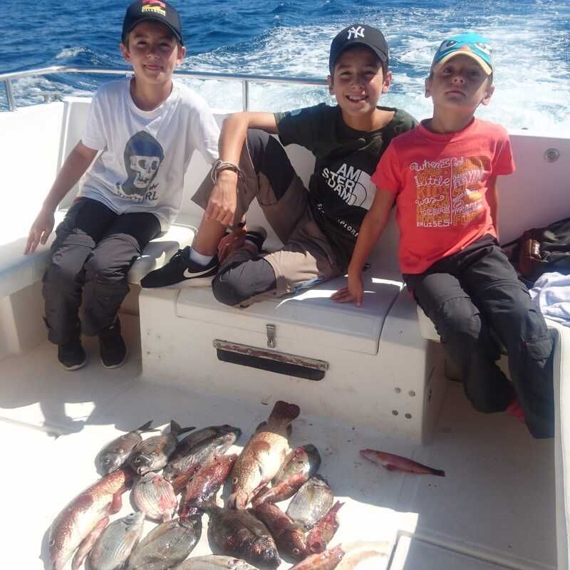 Tres niños están sentados en un bote con una buena pesca en la cubierta, con el océano de fondo.