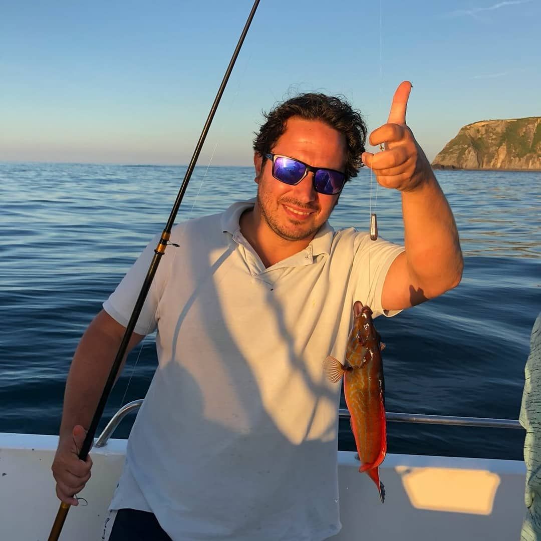 Un hombre en un bote sostiene un pez recién pescado mientras sonríe en el mar, cerca de una costa rocosa.