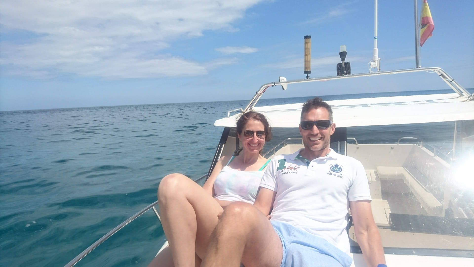 Dos personas se relajan en la cubierta de un barco sobre aguas azules y tranquilas bajo un cielo despejado.