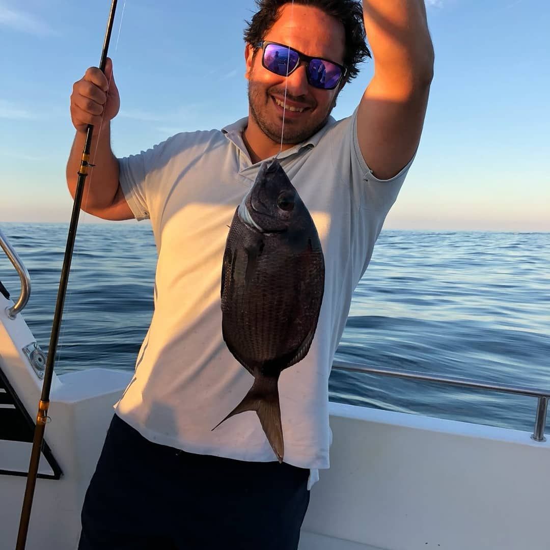 Un hombre en un bote sosteniendo un pez grande al atardecer, con gafas de sol, con el océano en calma de fondo.