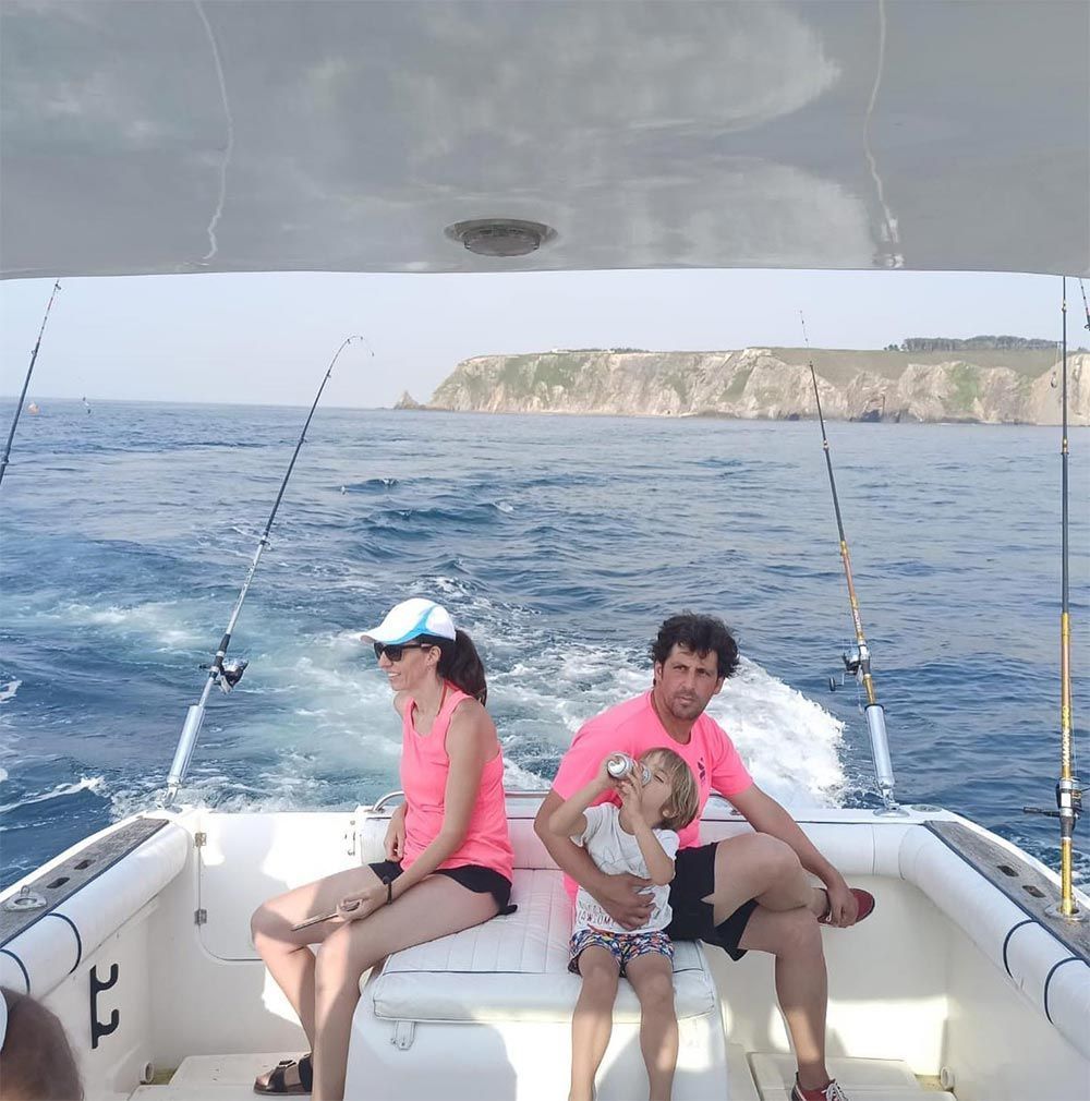 Tres personas relajándose en un barco, con cañas de pescar y una costa rocosa al fondo.
