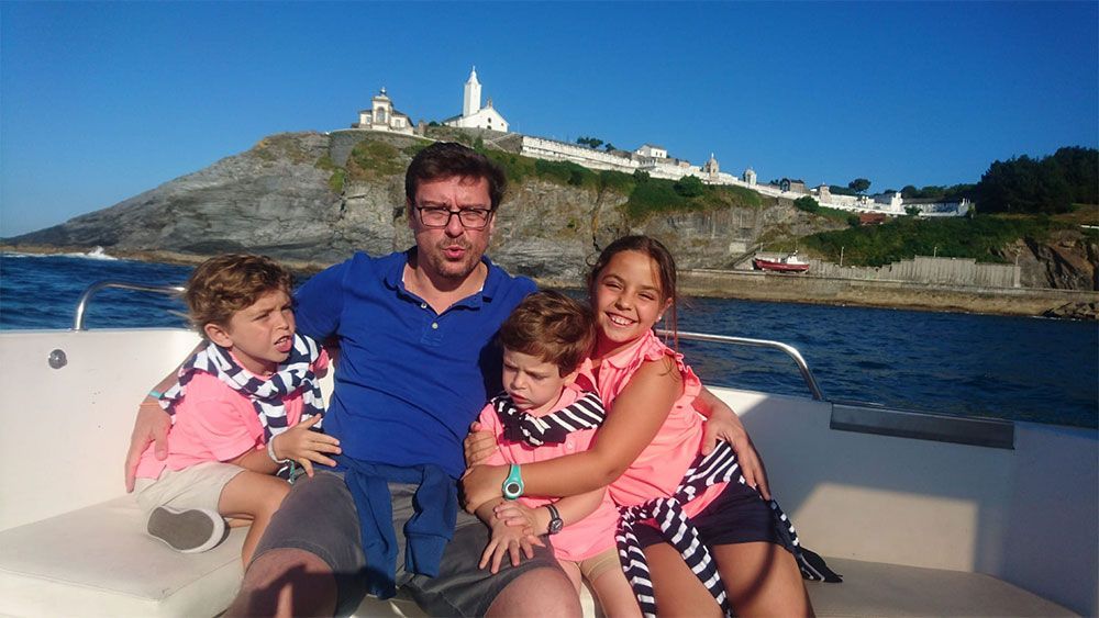 Familia sentada en un barco con un faro costero al fondo bajo un cielo azul.