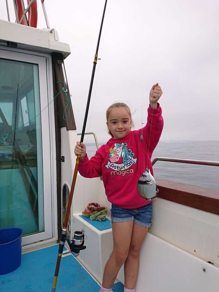 Una persona de pie en la cubierta de un barco, sujetando una cuerda y levantando un brazo, con agua gris y cielo nublado