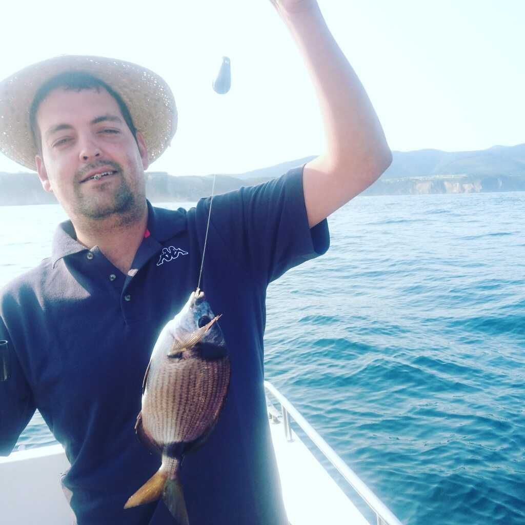 Un hombre en un bote sostiene un pez recién pescado sobre aguas azules con montañas al fondo.