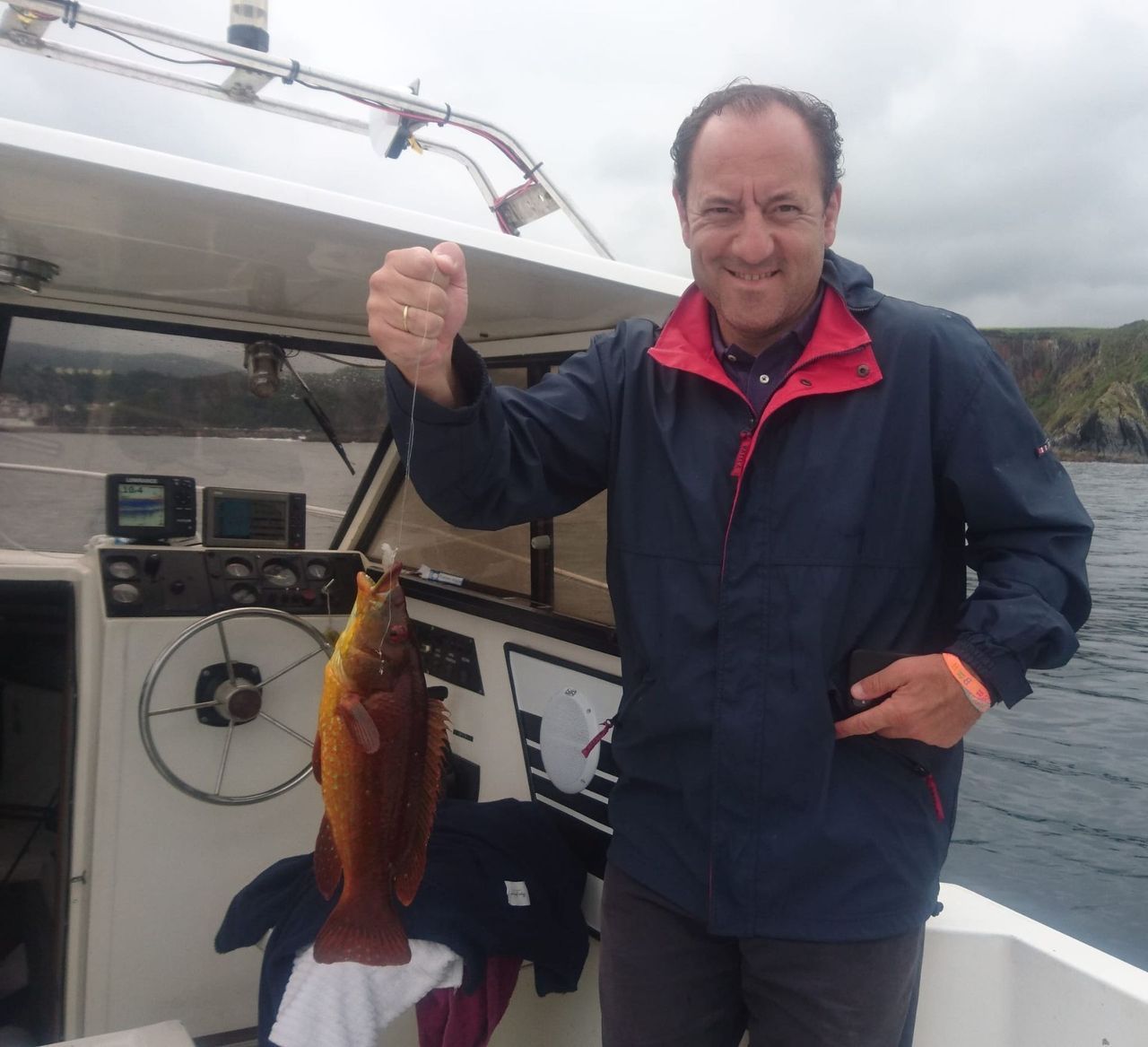 Un hombre en un bote sostiene un pez que ha pescado, con el agua turbia y la costa al fondo.