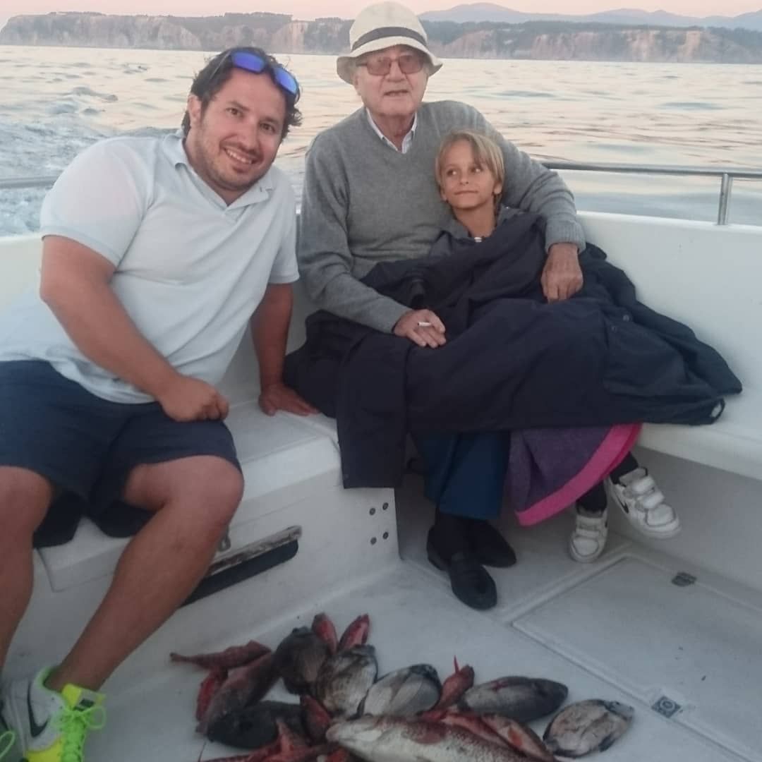 Tres personas en un bote con un montón de pescado en la cubierta, agua y acantilados al fondo.