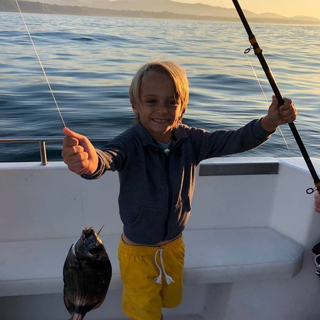 Un niño en un bote sosteniendo una caña de pescar y un pez al atardecer en aguas tranquilas.