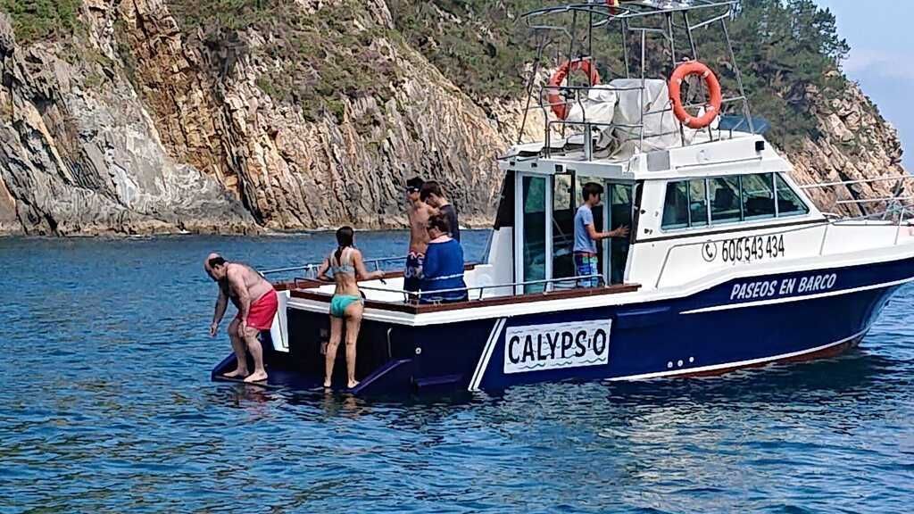 Personas subiendo a bordo de una pequeña embarcación blanca y azul cerca de una costa rocosa y arbolada.