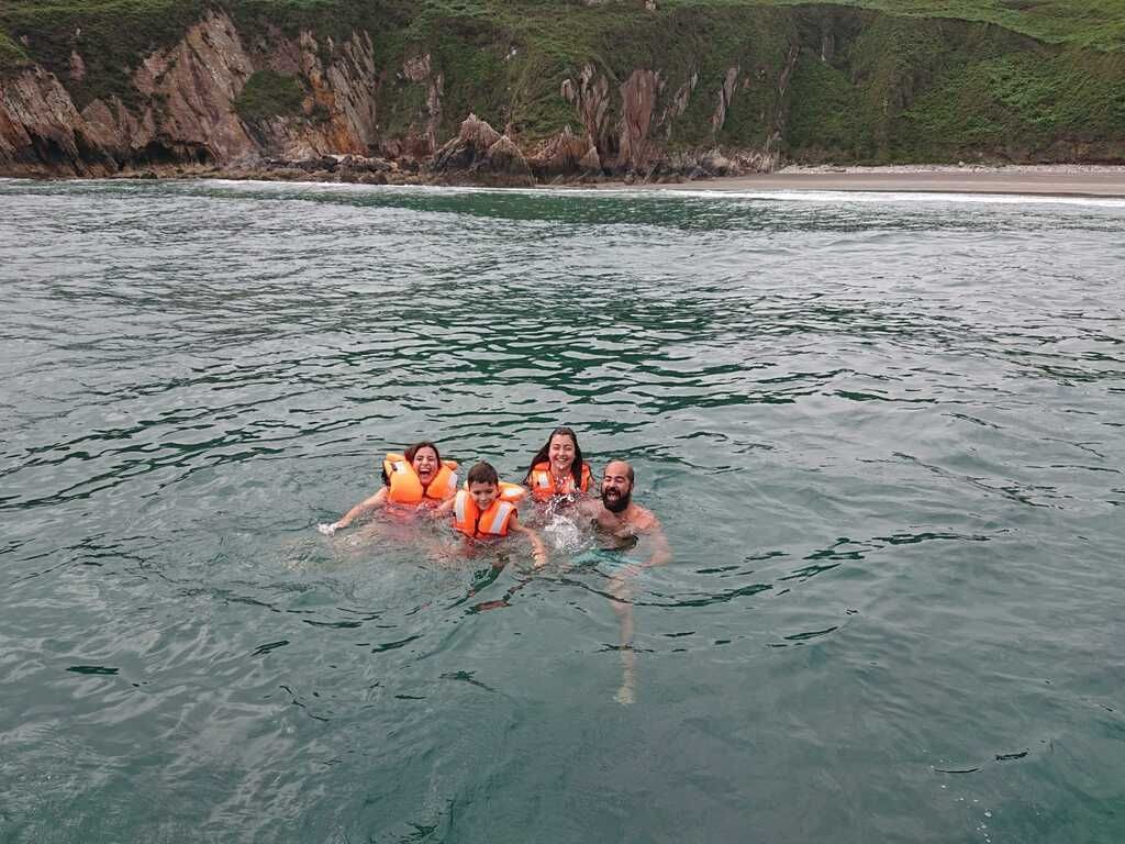 Cuatro personas con chalecos salvavidas naranjas flotando en el agua del océano cerca de una costa rocosa.