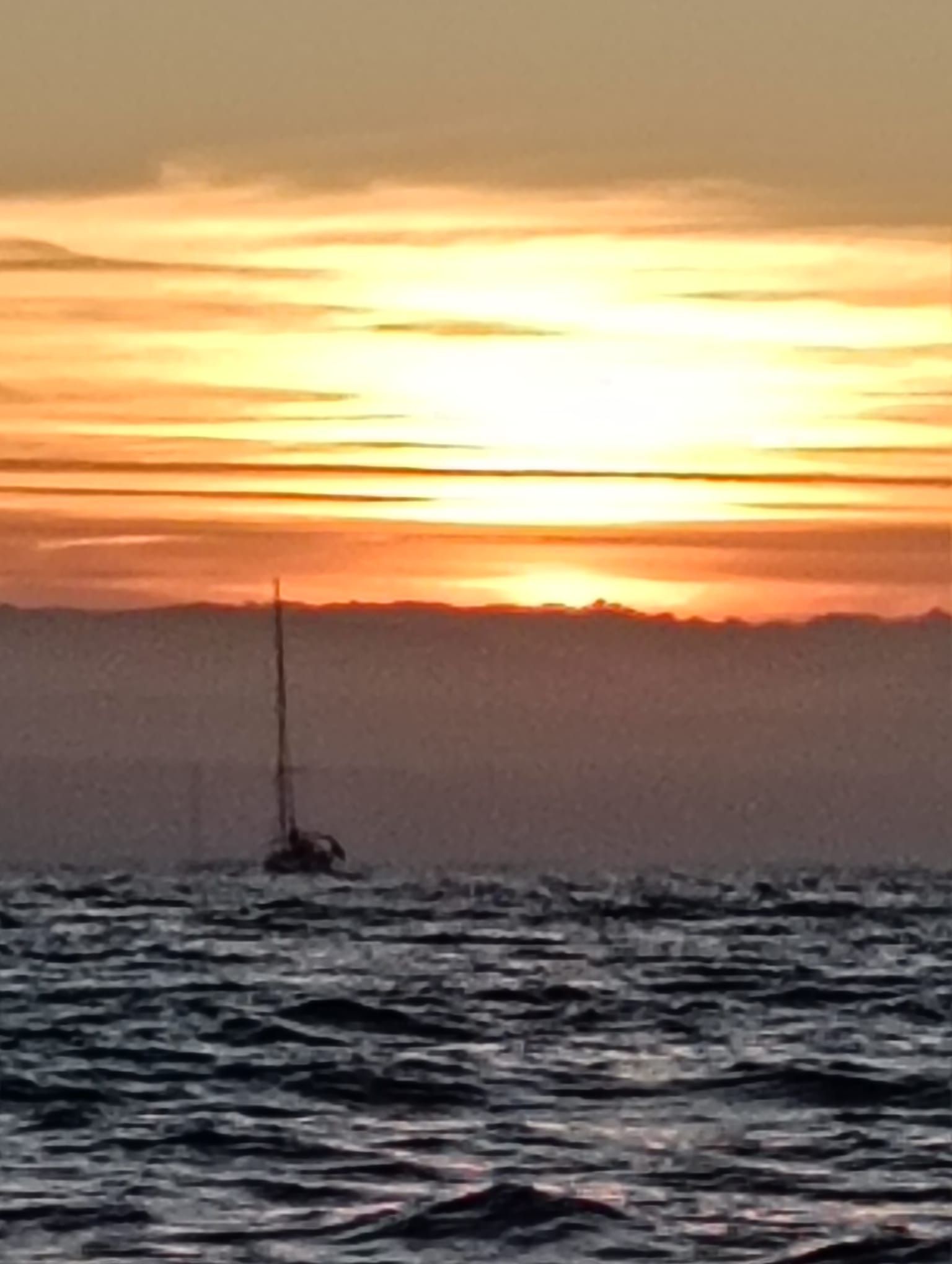 Silueta de un velero sobre aguas tranquilas al atardecer, con cielo anaranjado y nubes a lo lejos.