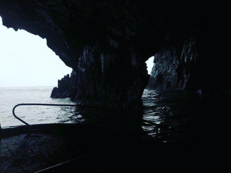 Una oscura cueva se abre sobre aguas tranquilas, con paredes rocosas y un horizonte luminoso más allá.