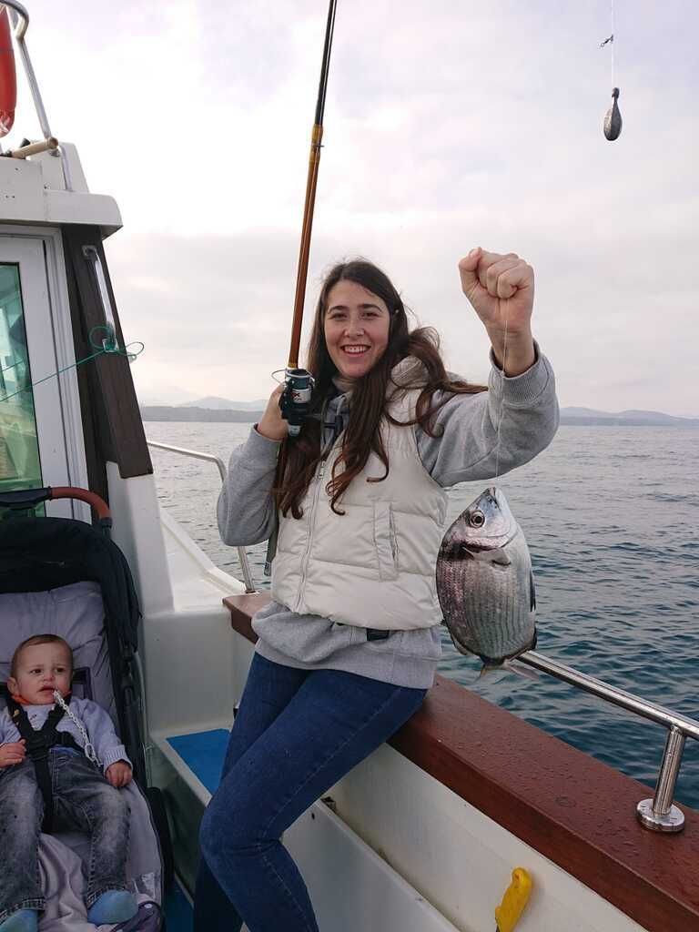 Una mujer con un chaleco blanco sonríe en un bote, sosteniendo un pez y levantando el puño, con un niño sentado cerca.