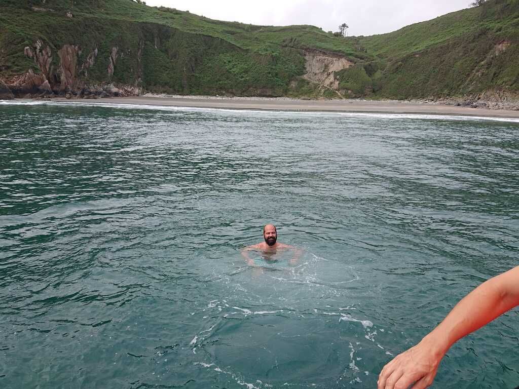 Persona nadando en aguas azules cerca de una costa rocosa y verde, con un brazo visible en primer plano.