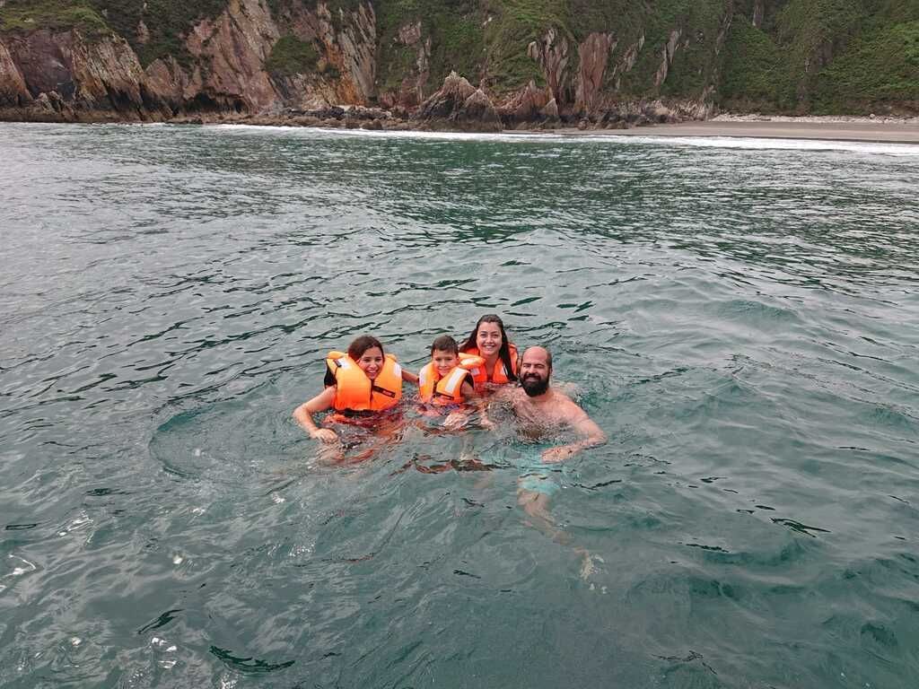 Tres personas con chalecos salvavidas naranjas flotando en aguas tranquilas cerca de una costa rocosa.
