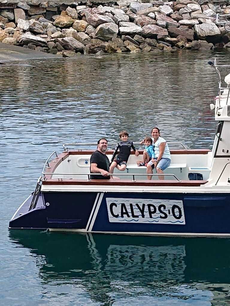 Personas sentadas en un barco llamado CALYPSO en aguas tranquilas cerca de una costa rocosa.