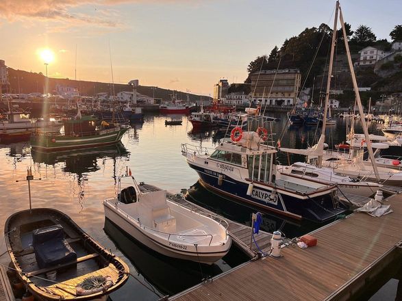 Puesta de sol sobre un puerto con varios barcos y yates amarrados junto a un paseo marítimo.