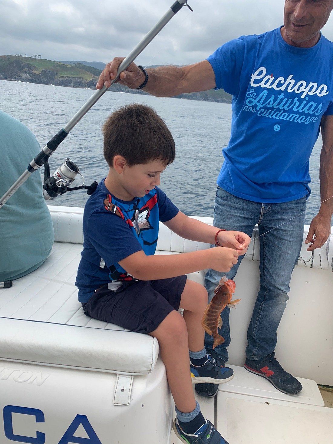 Un niño en un bote sostiene un pez mientras un adulto con una camisa azul permanece de pie cerca.