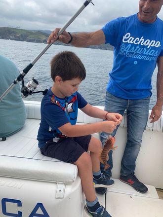 Un niño en un bote sostiene un pez mientras un adulto con una camisa azul permanece de pie cerca.
