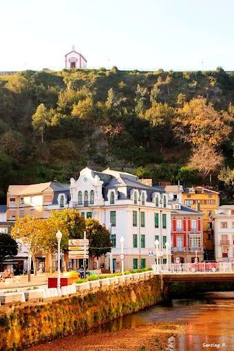 Edificios coloridos a orillas del río, bajo una colina boscosa, con una pequeña capilla en la cresta.