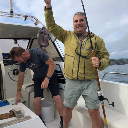 Dos hombres en un bote, sosteniendo cañas de pescar, con un pez colgando junto a ellos en la cubierta.
