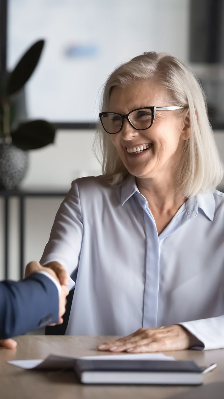 Une femme à lunettes serre la main d'une personne en souriant. À l'intérieur, chemise bleu clair, papiers sur la table.