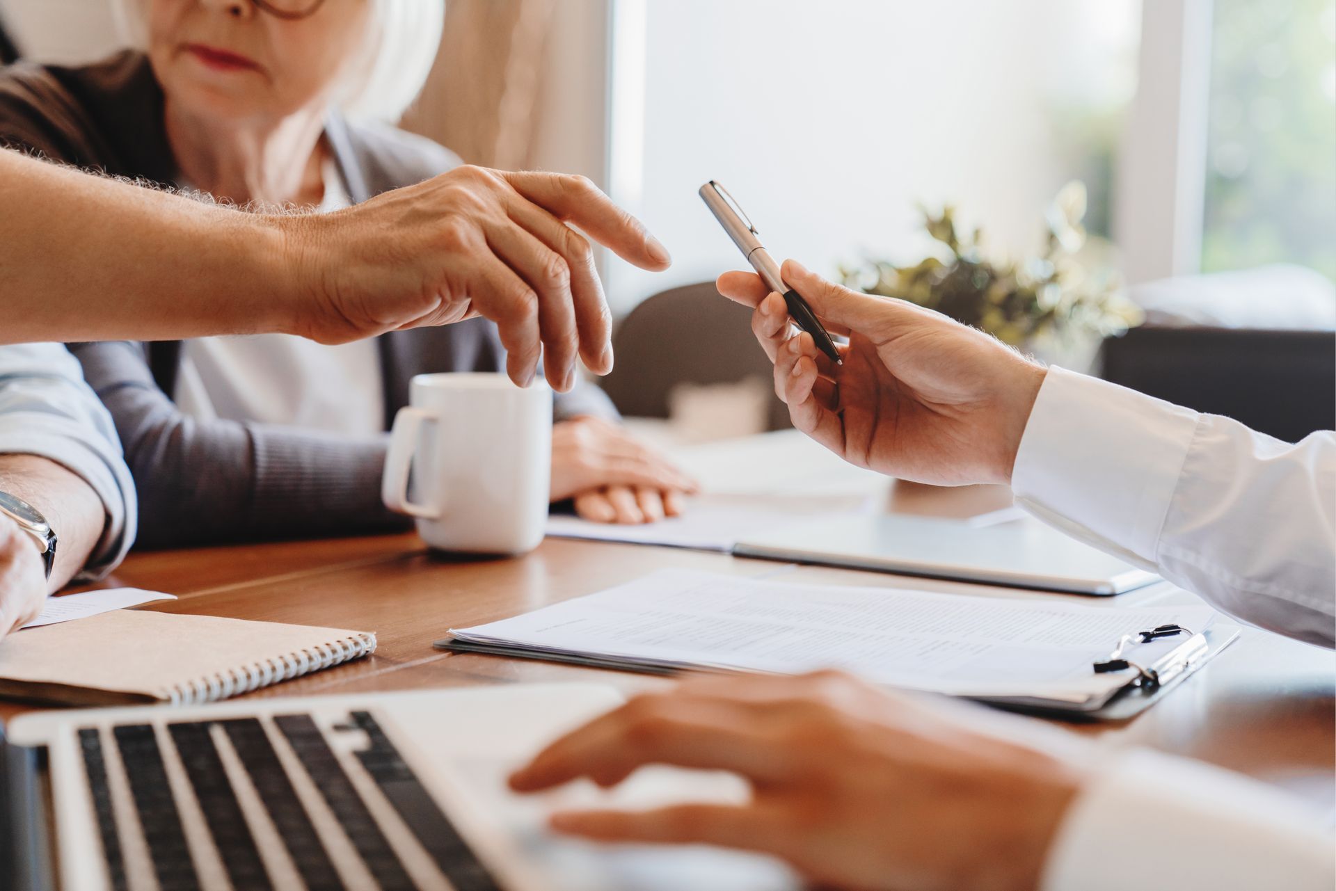 Une personne tend un stylo à quelqu'un assis à une table, probablement pour la signature de documents. Un ordinateur portable, une tasse de café et des papiers sont présents.