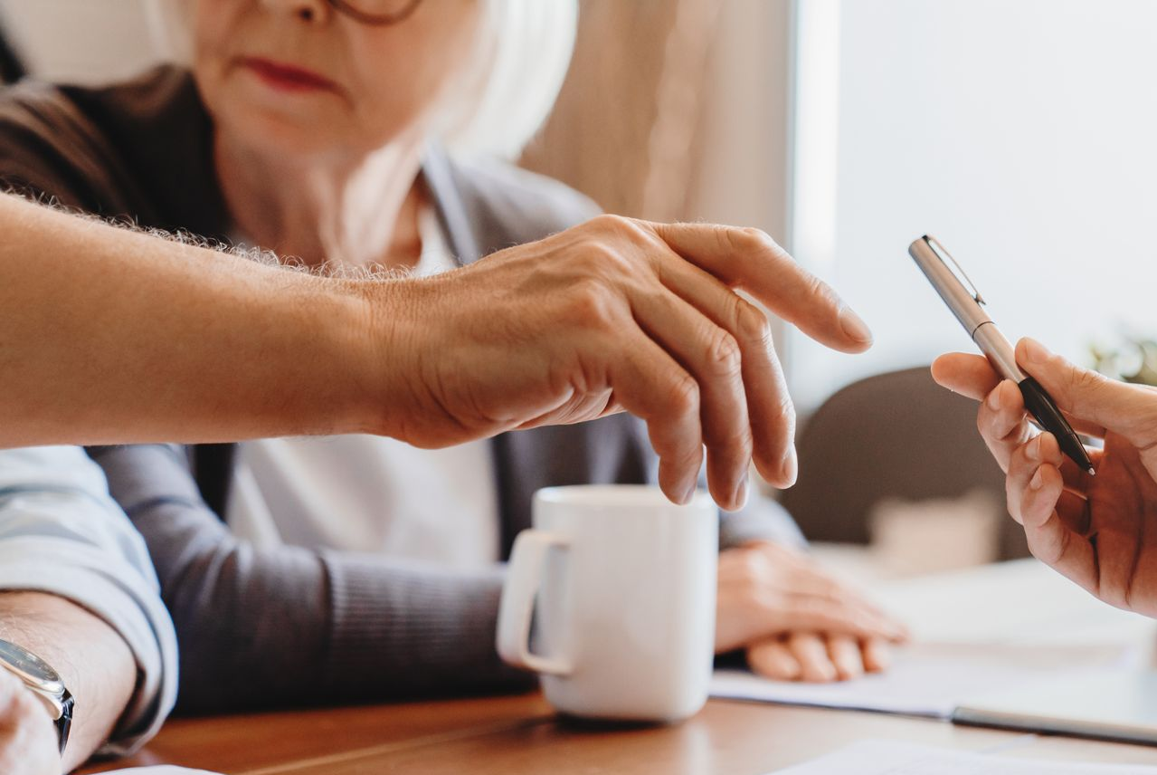 La main d'une personne tend vers un stylo tenu par une autre personne, assise à une table. Une tasse est présente.