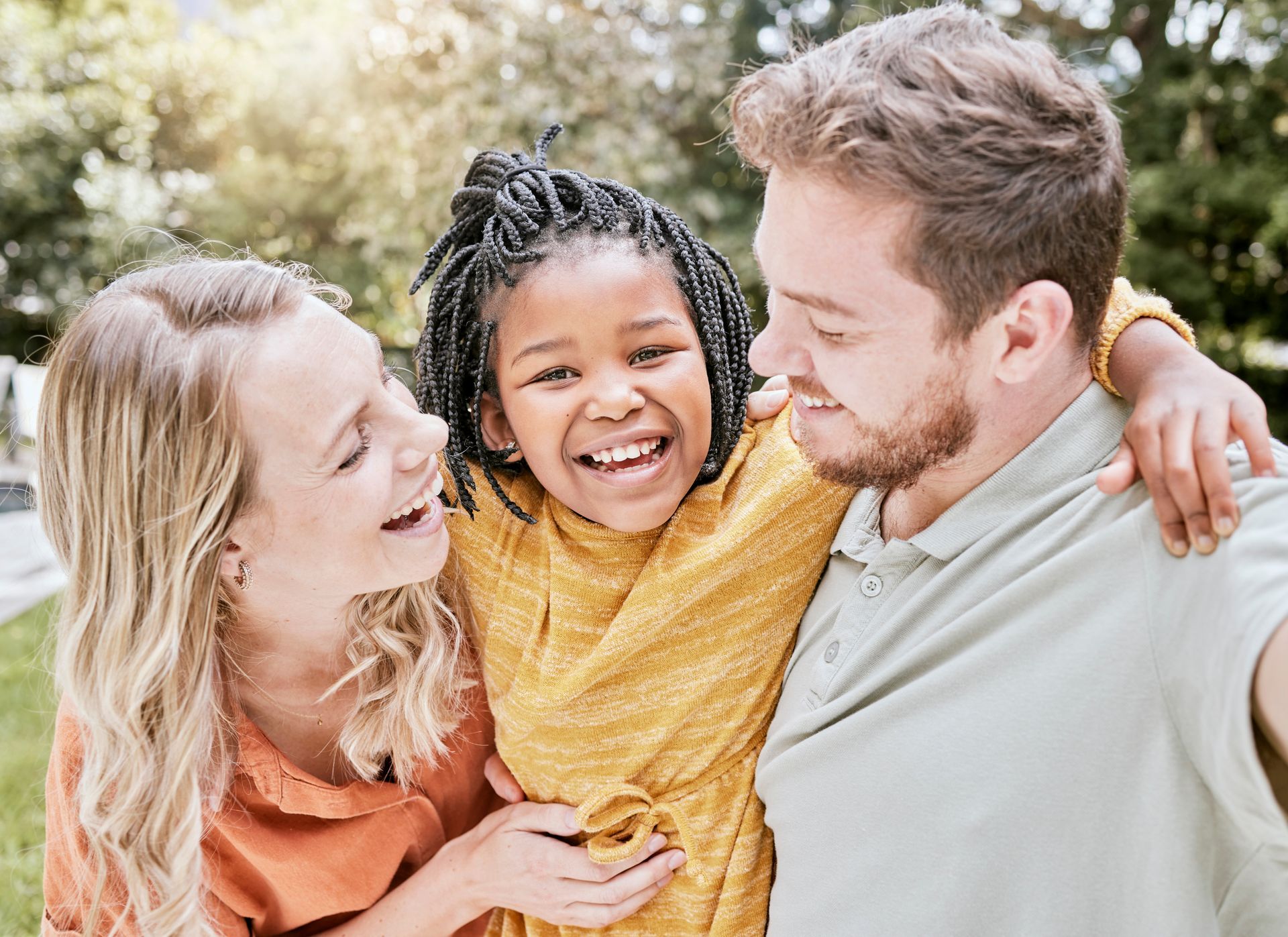 Une famille souriante s'enlaçant à l'extérieur : la fille porte un t-shirt jaune, les parents des t-shirts orange et gris.
