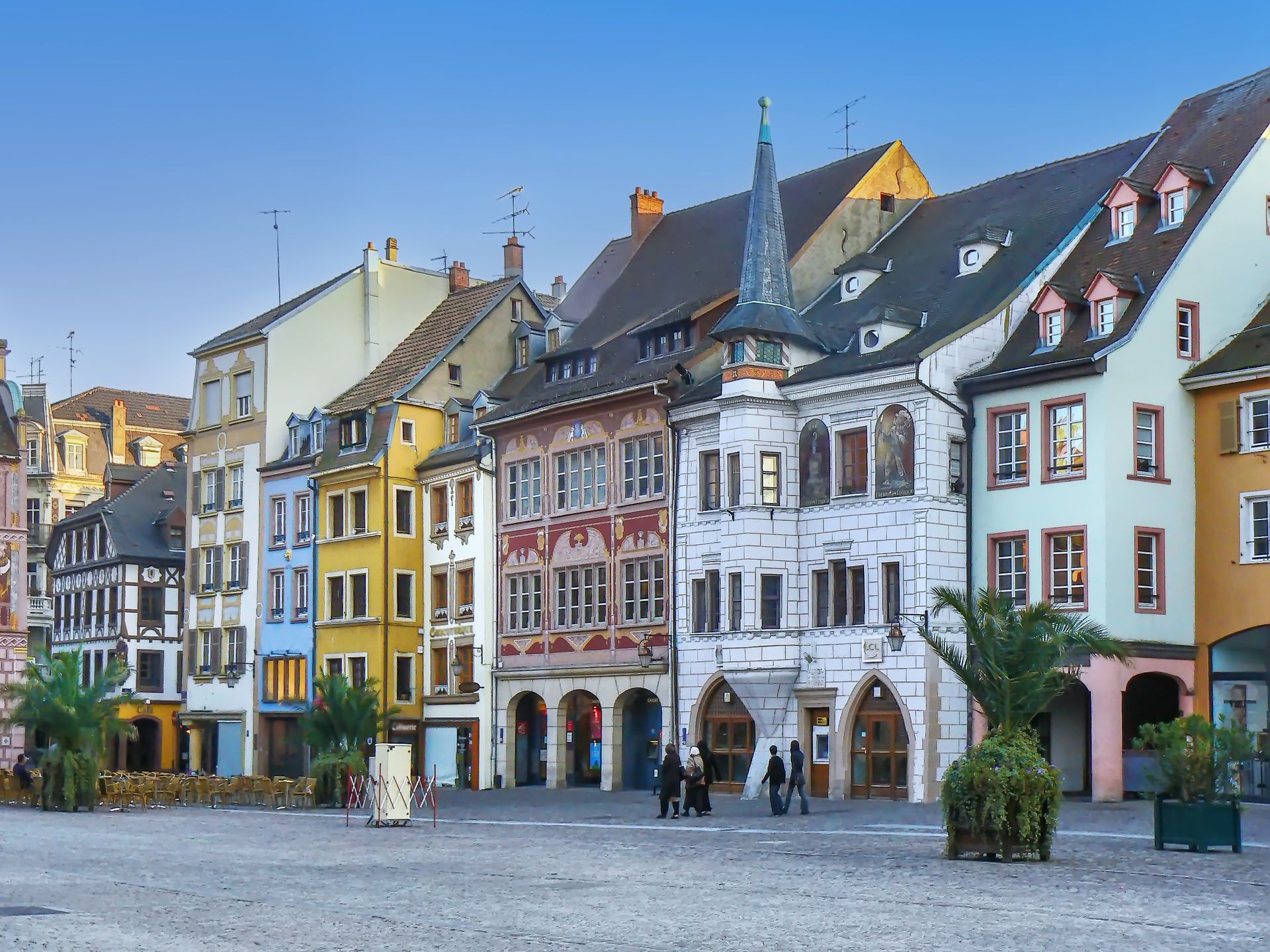 Des bâtiments historiques colorés bordent une place d'une ville européenne ; plusieurs personnes y passent.
