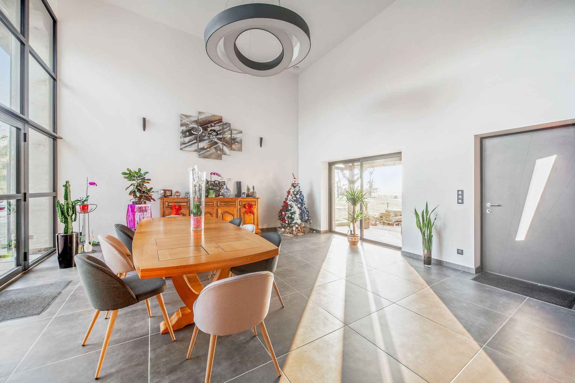 Salle à manger moderne et lumineuse avec table et chaises en bois, hautes fenêtres et plafonnier.