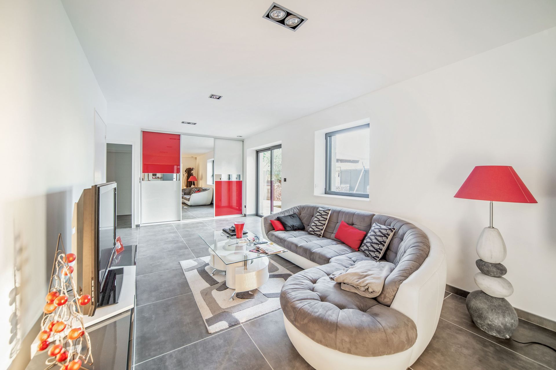 Salon moderne et lumineux avec canapé d'angle gris, touches de rouge, cheminée et grandes fenêtres