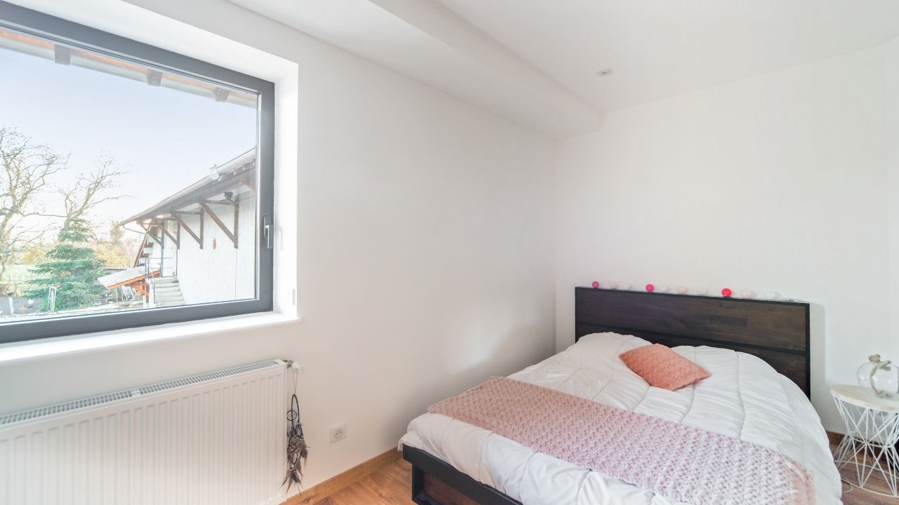 Chambre lumineuse avec un lit, des murs blancs, un parquet et une grande fenêtre