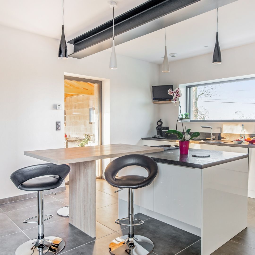 Cuisine blanche moderne avec îlot central, deux tabourets noirs et une grande fenêtre laissant entrer une lumière vive.