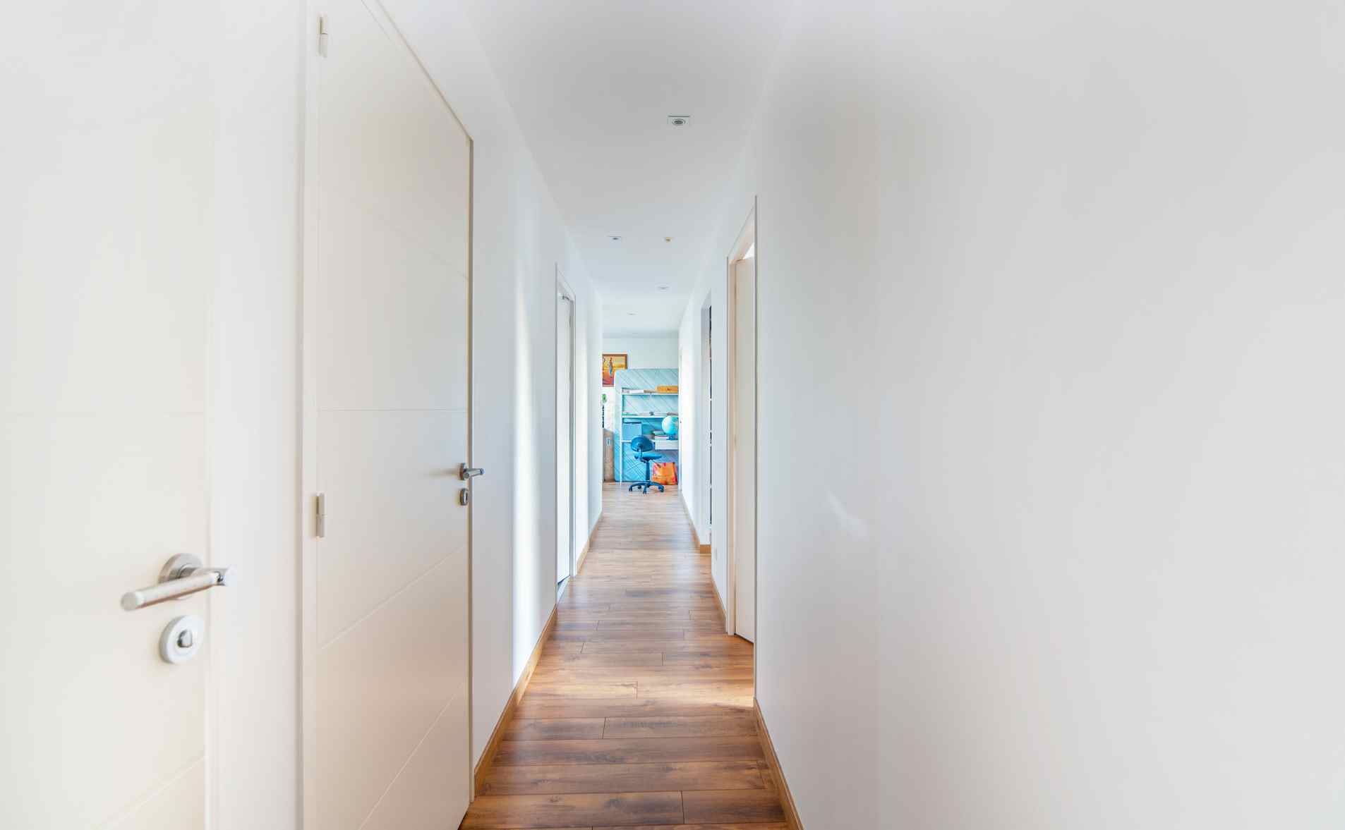 Un couloir blanc lumineux avec parquet et portes fermées mène à une pièce colorée
