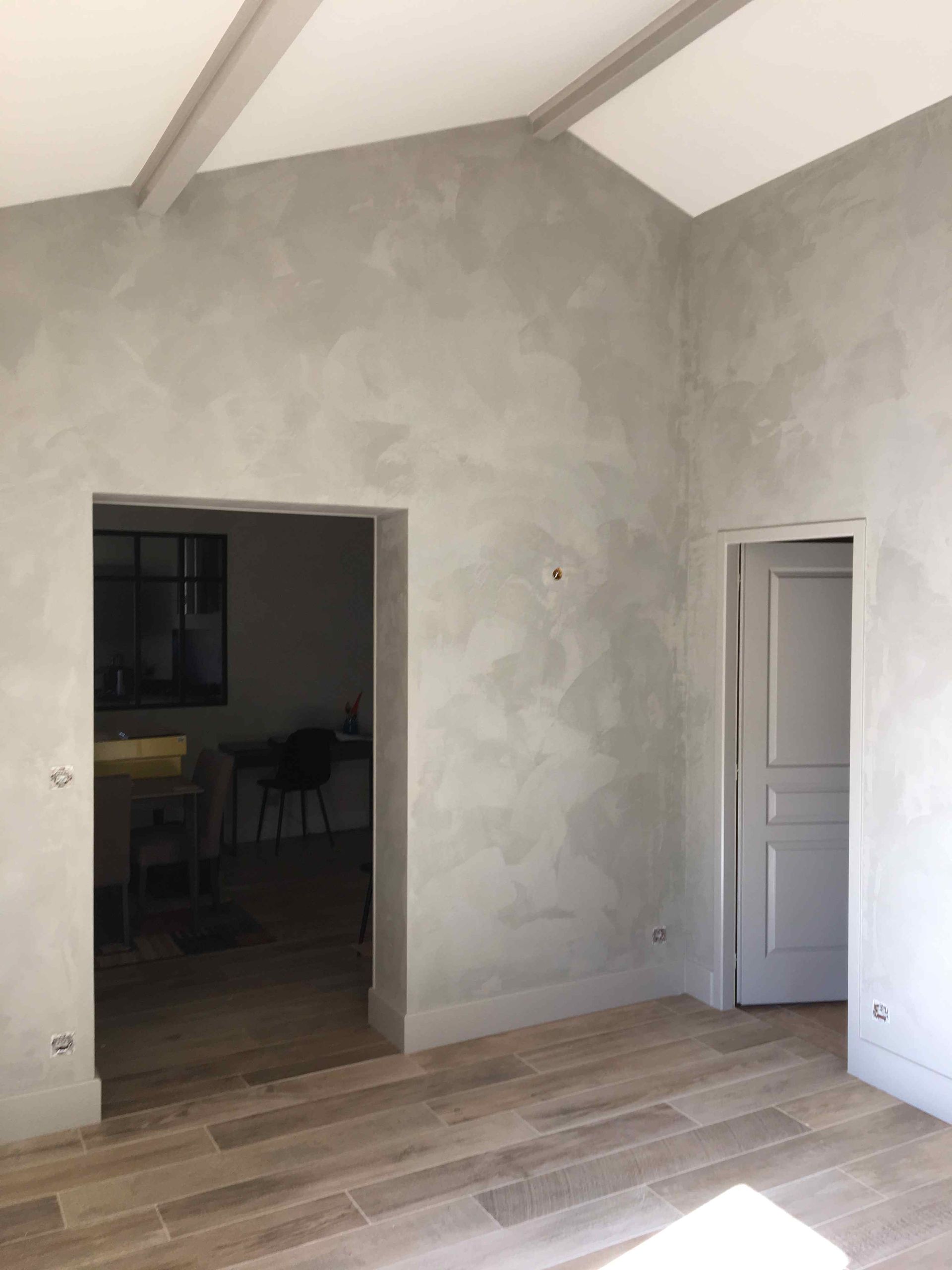 Pièce vide aux murs en plâtre gris, poutres apparentes au plafond, parquet et deux portes.
