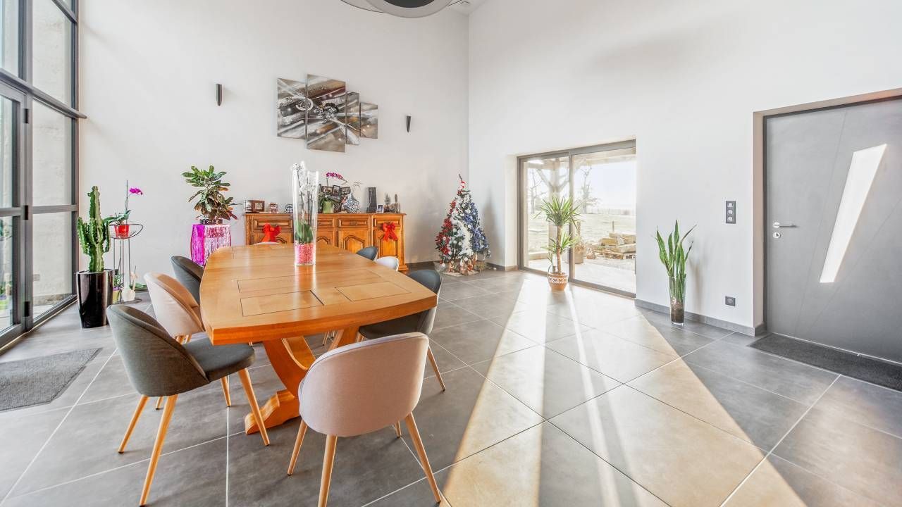 Salle à manger moderne avec table en bois, chaises, plantes en pot et lumière naturelle abondante grâce aux portes vitrées.