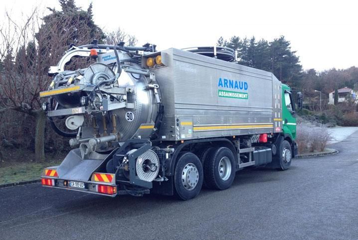 Un gros camion-benne vert et argenté, chargé de la collecte des déchets, est stationné dans une rue où le ciel est nuageux en arrière-plan.